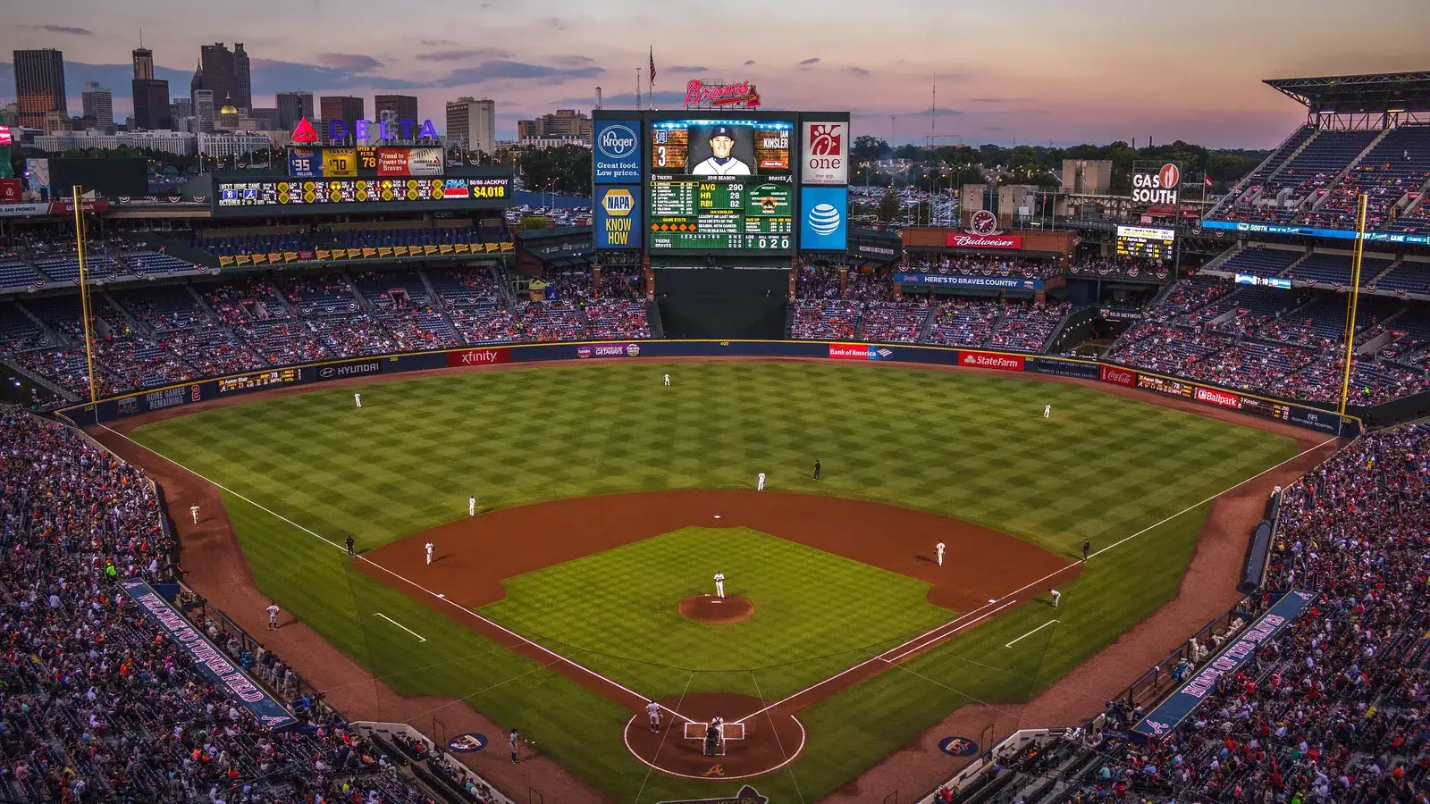 Baseball stadium at dusk seen from behind home plate, the city skyline beyond