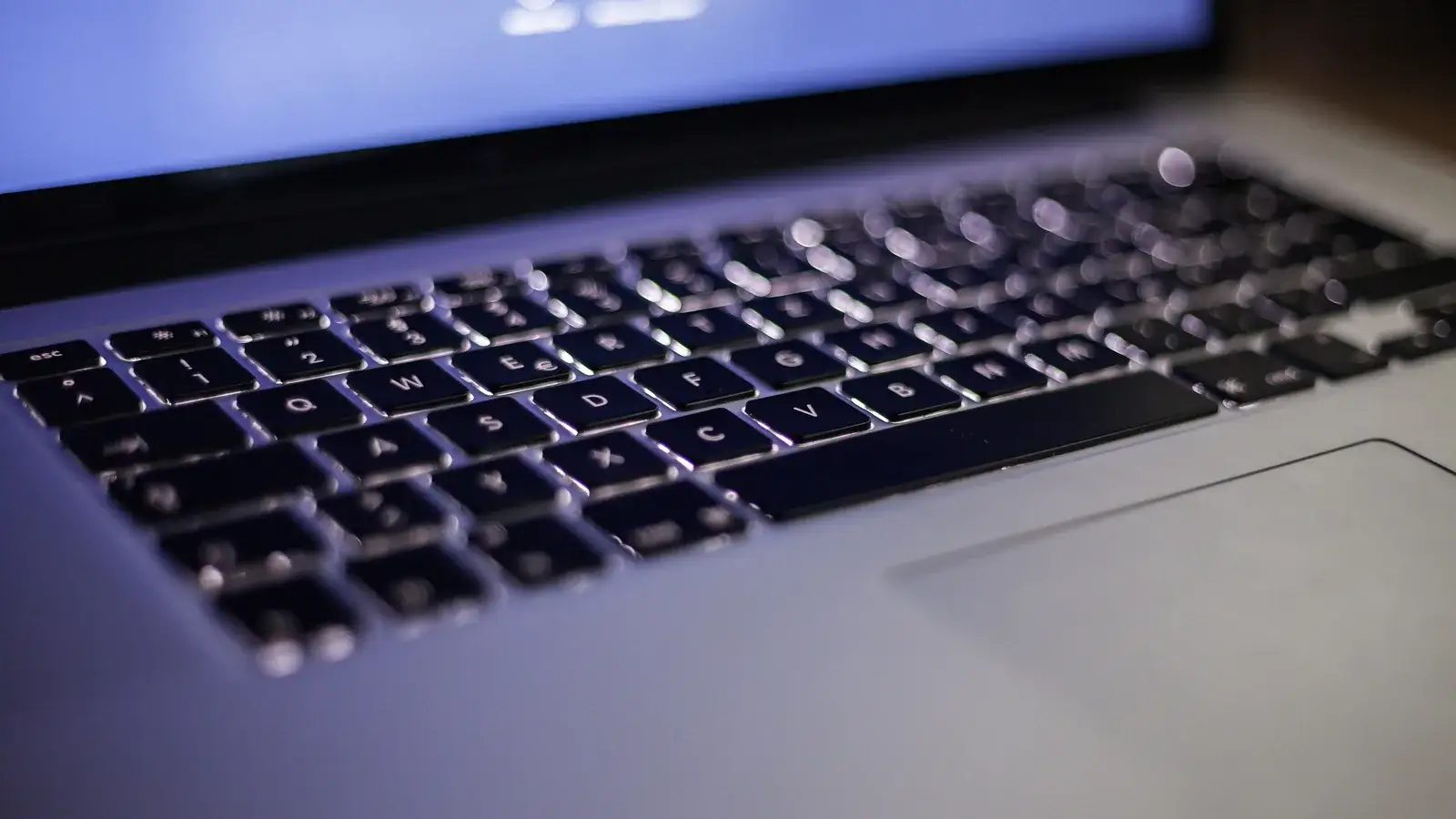 Backlit laptop keyboard glowing in a dark room, screen light above
