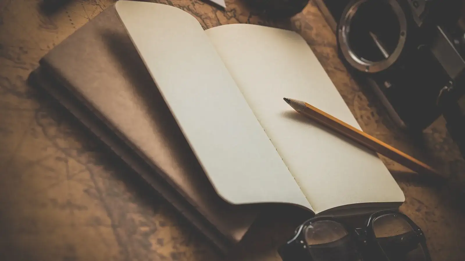 Open notebook and pencil on a dark desk with a camera and glasses