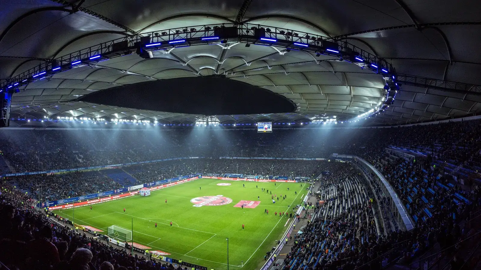 Floodlit football stadium at night seen from the upper stand, the pitch glowing under the roof lights