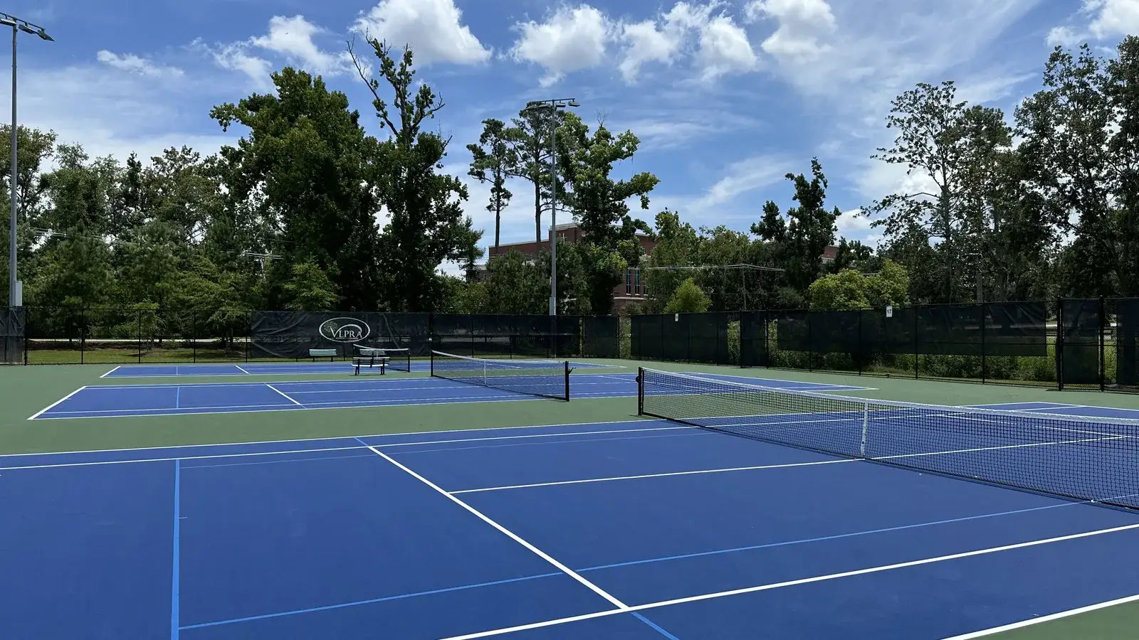 Row of blue hard tennis courts with nets and floodlight poles under a summer sky