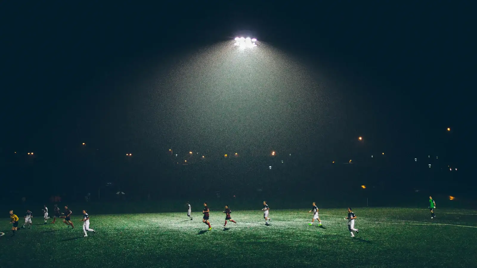 A single floodlight over a dark grass pitch at night with small figures playing football