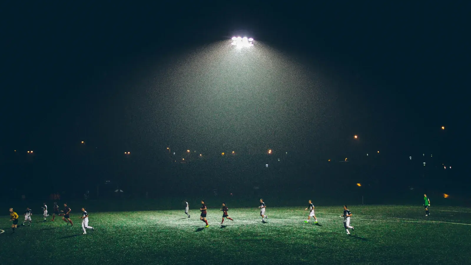 A single floodlight over a dark grass pitch at night with small figures playing football